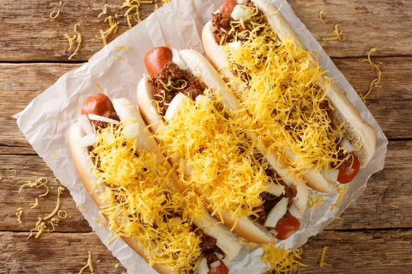 Beef hot dog in a bun covered with Cincinnati Chili, diced onions, and a mound of shredded cheddar cheese closeup in the paper on the table. Horizontal top view from abov