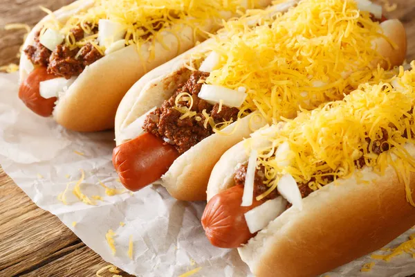 Beef hot dog in a bun covered with Cincinnati Chili, diced onions, and a mound of shredded cheddar cheese closeup in the paper on the table. Horizonta