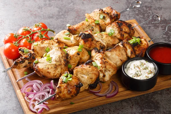 Delicious chicken kebabs on skewers with fresh vegetables and two sauces close-up on a wooden tray on a gray concrete background. horizonta