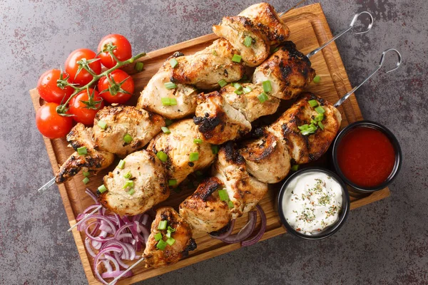 Delicious chicken kebabs on skewers with fresh vegetables and two sauces close-up on a wooden tray on a gray concrete background. horizontal top view from abov