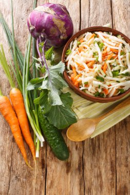 Tahta bir masadaki tabakta taze kohlrabi salatası, havuç, salatalık ve yeşil soğan. Yukarıdan dikey üst görünüm