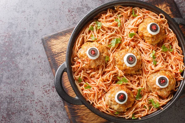 Halloween dinner of meatballs with monster eyes and pasta with tomato sauce in a frying pan on the table close-up. Horizontal top view from abov
