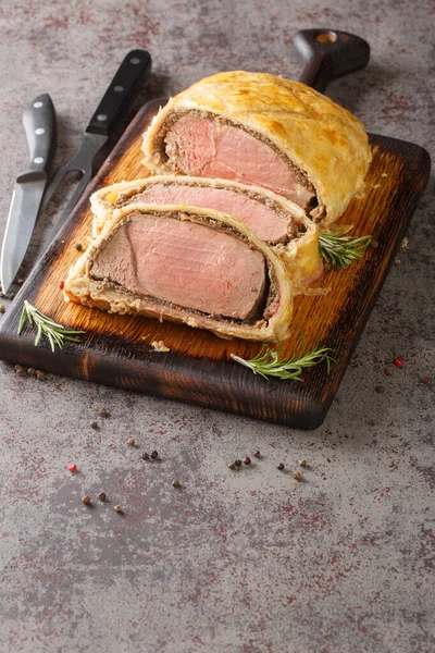 Sliced Beef Wellington on a wooden board with a sprig of rosemary on a grey background. Vertica