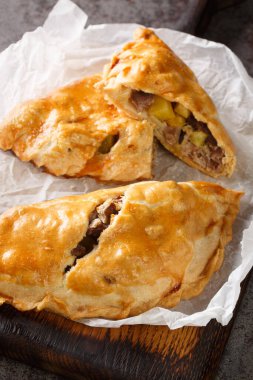 Traditional Cornish pasty filled with beef meat, potato and vegetables closeup on the paper on the wooden board. Vertica