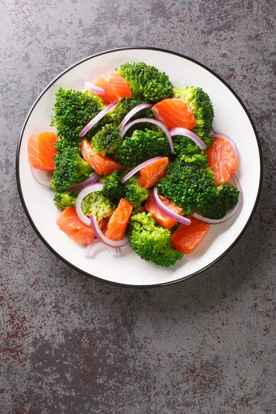 Dietary salad of steamed broccoli, salted salmon and red onion close-up in a plate on the table. Vertical top view from abov