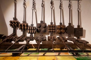 Eskiden waffle pişirmek için kullanılan yuvarlak kabuklu ve jantsız gibi çeşitli şekillerde eski metal waffle ızgaralarıyla dolu bir fırında bir duvar.