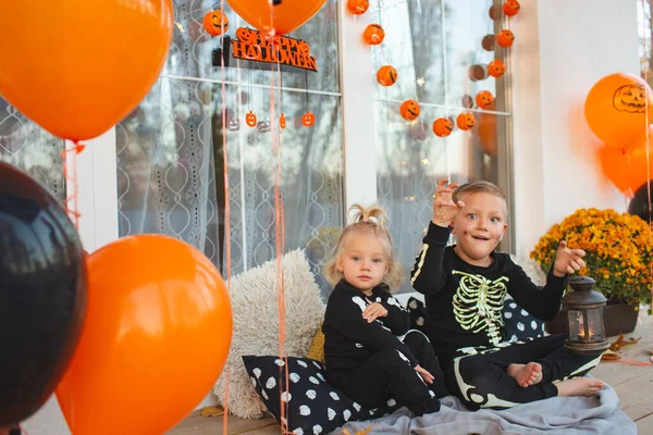 Kids dressed as skeletons  play on the terrace of their house. The space is decorated with themed balloons, garlands, chrysanthemums. Halloween decorations