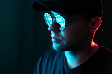 Gözlüklü ve suratında neon ışığı olan bir adam. Stüdyonun karanlık portresindeki ilan panosundan mavi ve kırmızı renkler. Güneş gözlüğündeki yansıma. Dramatik şehir gece portresindeki siyah arkaplan. Siber teknoloji.