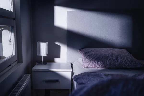 Bed at night in dark bedroom. Blue light and moonlight from window. Pillow, sheet and duvet ready for sleeping. Bedside table and nightstand. Scandinavian home interior design. Scary shade and shadow.