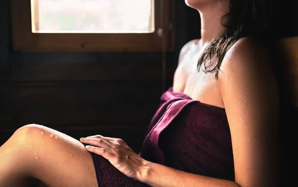 Woman in sauna. Sweat on skin, towel on body and wet hair. Steam room bath in relax spa in Finland in summer. Traditional Finnish lifestyle. Wellness and warm rest therapy in dark wood home interior.