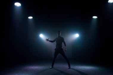 Günlük kıyafetli bir adam karanlık bir sahnede dans pozu verir. Arka ışıklandırma sahne ışığı. Bir dansçının silüeti..