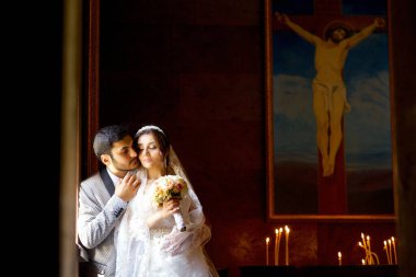 Düğün töreninden sonra kilisede duran genç ve güzel bir çift. Beyaz elbiseli kız gelin. Açık renk ceketli bir damat. Arka planda İsa 'nın ikonu.