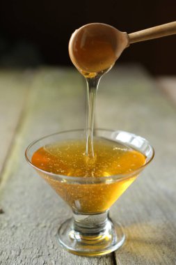 Young light floral honey is poured into a glass vase. The vase stands on the wooden surface of the table. C wooden spoon pouring honey in a bowl.