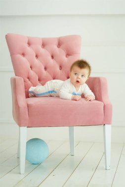 A baby boy is sitting on a pink chair. Next to it is a blue ball. Basic colors for children of different genders.