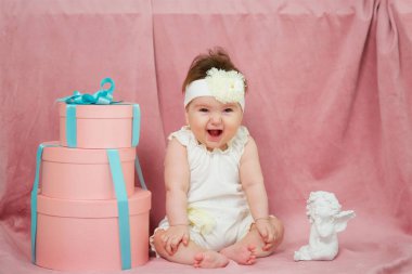 A little girl sitting on a pink background in white clothes. Next big gift and a statue of an angel.