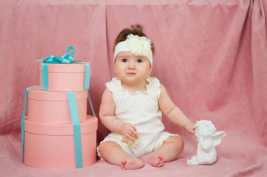 A little girl sitting on a pink background in white clothes. Next big gift and a statue of an angel.