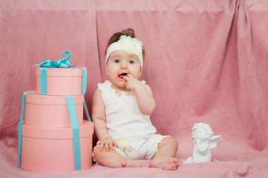A little girl sitting on a pink background in white clothes. Next big gift and a statue of an angel.