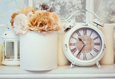 İçi şamdanlar, mumlar, çiçekler gibi diğer eşyalarla çevrili bir rafta çevirmeli çalar saati olan antika bir saat..