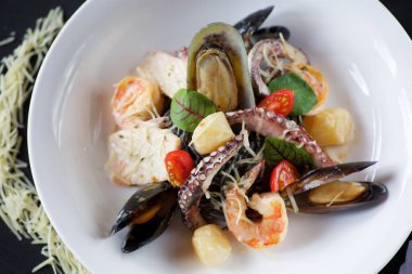 Üst manzara - deniz ürünleri - karides midyeleri ve ahtapotlar, siyah bir yüzey üzerinde beyaz bir tabakta peynir ve fesleğen yapraklarıyla servis edilir.