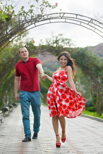 緑と新鮮な花のアーチで公園を歩いている男と女の子の若いカップル — ストック写真