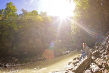 Yetişkin bir kız baharda ya da sonbaharda bir dağ nehrinin kıyısında bir kayanın üzerinde oturur. Vahşi doğada mahremiyetin tadını çıkar. Işıklı güneş.