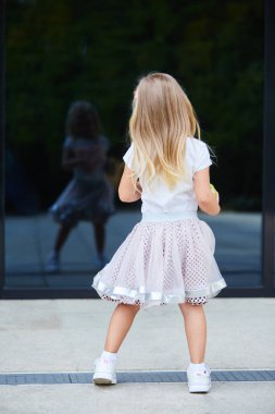 Beyaz tişörtlü ve pembe etekli küçük sarışın bir kız cam yüzeydeki yansımasına bakıyor. Çocukluk ve yetişkin taklidi kavramı..