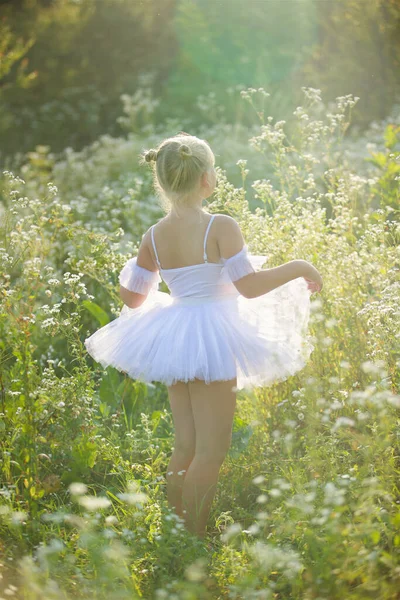 白いバレエのチュチュのブロンドの女の子が背の高いハーブや花の間に立っています カメラに背を向けた 完全な成長で — ストック写真