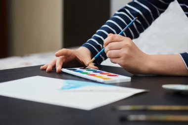 Mavi bir fırçayla çocukların elleri ahşap bir masanın üzerine suluboya bir resim çiziyor. Yan görünüm.