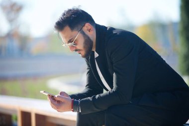 Başarılı bir temsilci, modern binanın önündeki bankta oturan sakallı, zengin ve modaya uygun bir adam. Erkekler için güneş gözlüğü.