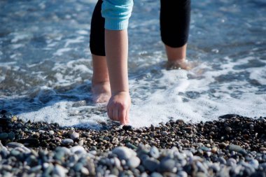 Cinsiyeti belirsiz bir adamın eli deniz ya da okyanus kıyısında taş toplar..