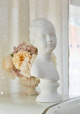 Alçıdan yapılmış bir kızın heykeli, büst. Arka planda vazoda çiçekler var. Vintage iç mimari.
