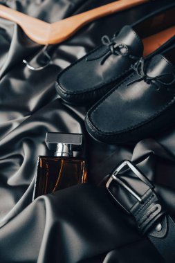 Erkek aksesuarları. Erkek gardırobunun ayrıntıları. Bir şişe erkek parfümü, deri ayakkabılar ve kemer saten kumaşın üzerinde..