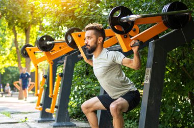 Spor salonu aç. Atletik adam dışarıda eğitim makinesiyle çömeliyor. Belediye spor ekipmanları üzerine sokak eğitimi. 