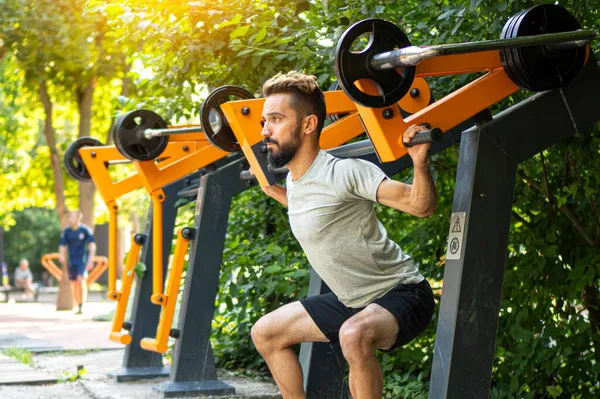 Spor salonu aç. Atletik adam dışarıda eğitim makinesiyle çömeliyor. Belediye spor ekipmanları üzerine sokak eğitimi. 