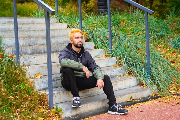 秋の公園の階段に座ってブロンドで染め髪のスタイリッシュなスポーツマン — ストック写真