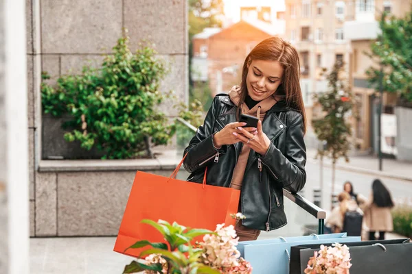年轻女子在外面的社交媒体上通过电话聊天 她旁边有五颜六色的购物袋 快乐的生活方式 假日销售的概念 — 图库照片