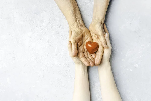 Taking care for the elderly concept with a wrinkled hands and young hands holding a red heart symbol.
