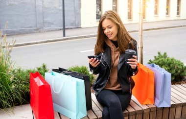 Beautiful young woman sitting with shopping bags on a bench. She is wearing casual style. In one hand she holds coffee. In other hand she holds cellphone.