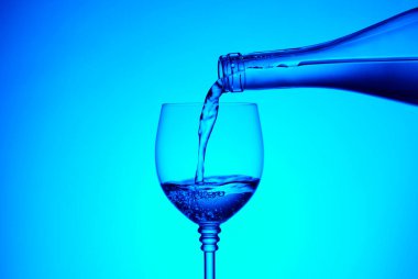 Cold water pouring into the glass from the glass bottle on a blue background. The concept of quality drinking water.