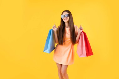 Fashion woman wearing summer dress and sunglasses holding shopping bags on a yellow background with copy space. Holidays, summer sales, discounts, shopping concept.