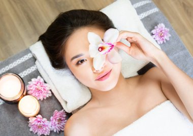 Day at spa. Beautiful asian lady having spa, massage and aromatherapy session. Pretty korean young woman holding orchid flower on her face, while laying next to the flowers, and candle. Relaxation atmosphere.