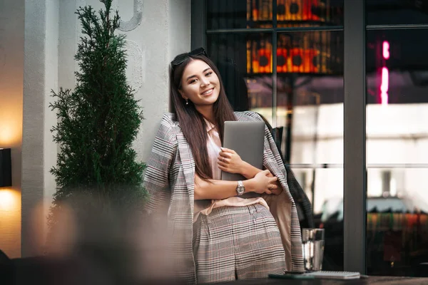 Young asian business woman with laptop in her hands looking at us and posing near the big window. Freelance, business online, busy lifestyle concept. Cozy street atmosphere.