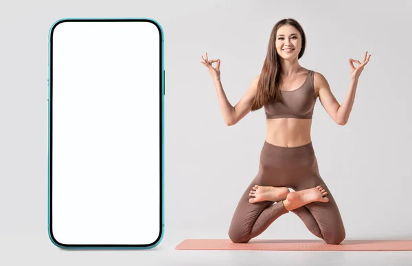 Yoga or fitness application blank. Fit asian woman practicing yoga, keeping balance, while standing on a knees in difficult lotus pose near the modern smartphone with empty screen. Healthy lifestyle, yoga, stretching, fitness concept.