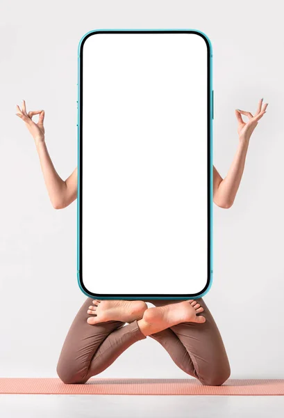 Yoga or fitness creative application blank for your design. Fit woman practicing yoga, keeping balance, while standing on a knees in a difficult lotus pose and holding modern smartphone with empty white screen. Vertical.