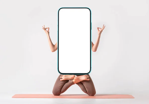 Yoga or fitness creative application blank for your design. Fit woman practicing yoga, keeping balance, while standing on a knees in a difficult lotus pose and holding modern smartphone with empty white screen.