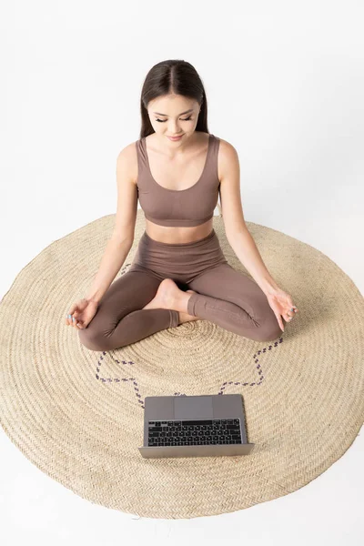 Online yoga lessons. Asian woman sitting in lotus pose against the laptop. Vertical photography.