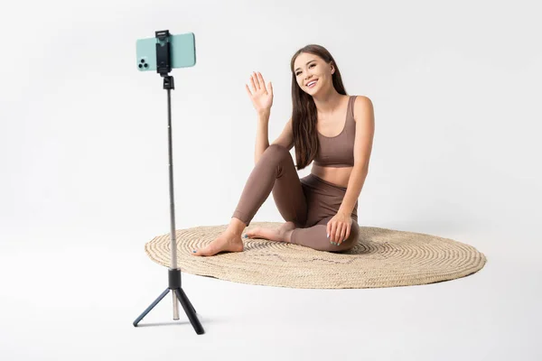Yoga online. Online training at home with fit asian woman welcoming audience through the cellphone holding on tripod.