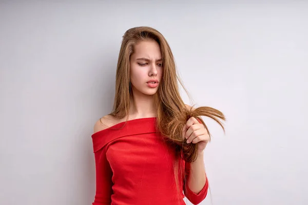 Δυστυχισμένη νεαρή κοπέλα δυσαρεστημένη από τα ξηρά μαλλιά με σχισμές άκρες, ενοχλημένη, απομονωμένη στα λευκά — Φωτογραφία Αρχείου