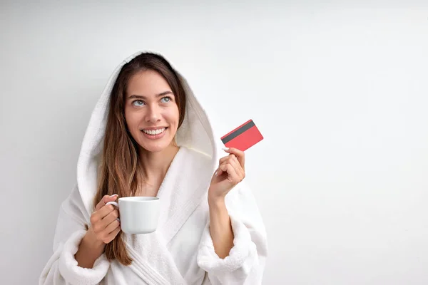 Mujer joven positiva que usa albornoz en el spa de bienestar con tarjeta de crédito, aislada en blanco —  Fotos de Stock