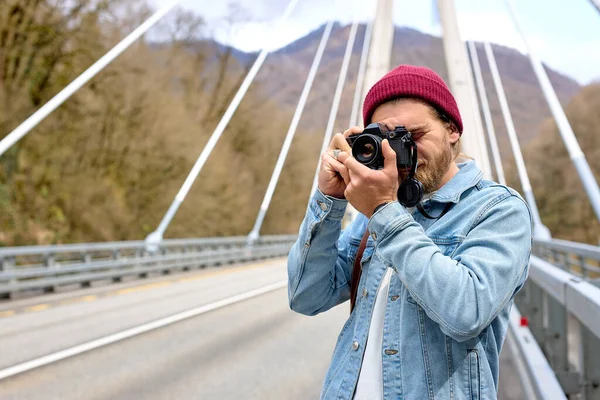 Hipster macho em jaqueta de ganga na ponte no campo usando câmera — Fotografia de Stock
